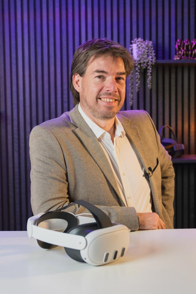 Dirk Koke in Geschäftskleidung mit einem lächelnden Gesichtsausdruck sitzt vor einem violetten Hintergrund und hält eine VR-Brille in der Hand.